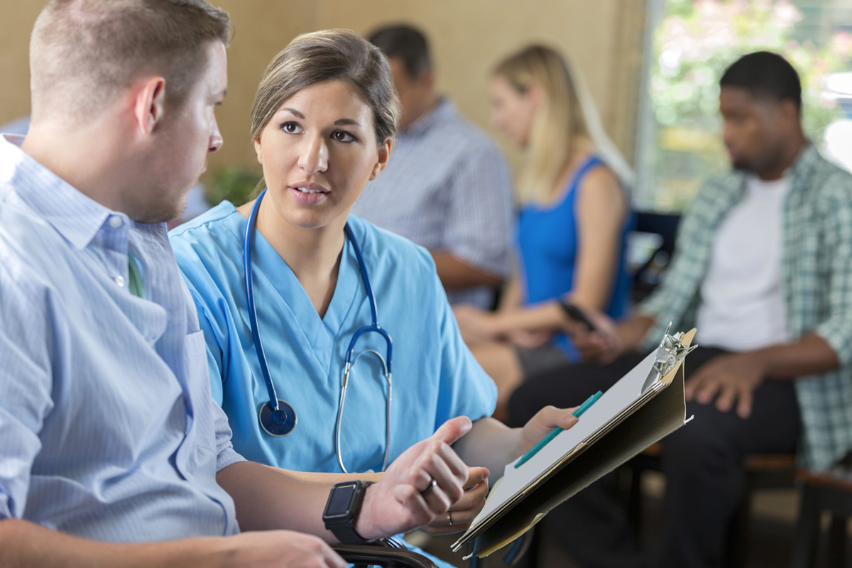 Mid adult Caucasian woman is nurse in hospital emergency room triage center. She is examining and talking with a patient, while other patient's wait behind them in waiting room. Nurse is reviewing paperwork with male patient.